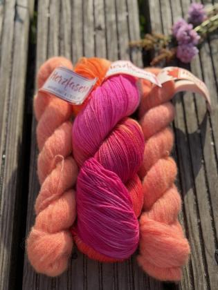 Handgefärbtes Petit-Provence-Garnset „Sonnenkuss“ von Herzfasern. In der Mitte liegt ein Strang Herzfasern No. 11 „Petras Farben 2.0“ in leuchtendem Pink, Neonpink und Orange. Links und rechts befinden sich pfirsichfarbene Stränge Herzfasern No. 93 aus Ka